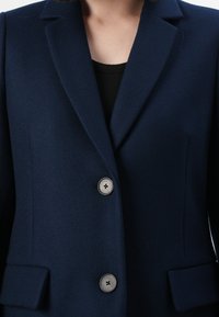 Dunkelblauer taillierter Blazer mit zwei silbernen Knöpfen und aufgesetzten Taschen, getragen über einem schwarzen gerippten Oberteil.
