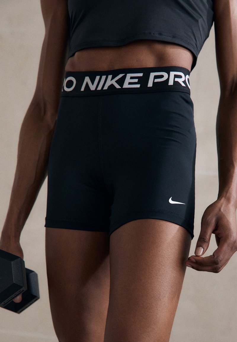 Personne portant un short Nike Pro noir et un crop top noir tenant un haltère noir dans une main, sur un fond neutre.