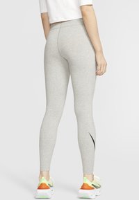 Šedé legíny s vysokým pasem vyrobené z elastického materiálu, s černým logem swoosh na levé noze, doplněné barevnými teniskami.