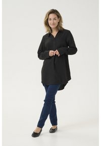 Schwarze Tunika mit langen Ärmeln und kragenfreiem Ausschnitt, aus strukturiertem Stoff. Kombination mit dunkelblauen Slim-Fit-Hosen und schwarzen Ballerinas.