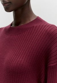 Pull en maille côtelée bordeaux avec un col rond ; présente un tissu texturé avec des rayures verticales, offrant un aspect doux et chaud.