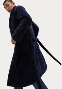 Granatowy, długi, pluszowy szlafrok o teksturowanej powierzchni, z szerokimi rękawami, paskiem oraz bocznymi kieszeniami. Model pozuje z jedną ręką w kieszeni.