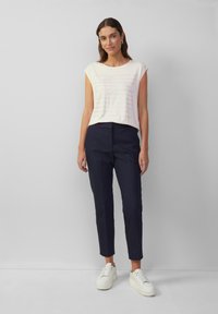 Cremefarbene kurzärmelige Bluse mit strukturierten Linien und Textdesign, kombiniert mit marineblauen schmalen Hosen und weißen Sneakern, vor einem einfarbigen Hintergrund.