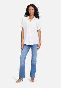 Weißes kurzärmliges Hemd mit Knöpfen, aus strukturiertem Stoff. Kombiniert mit hellblauen, ausgestellt geschnittenen Jeans und beigen, mit Absatz versehenen Sandalen. Einfaches Design.