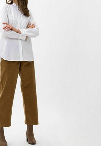 Biała koszula z długim rękawem i zapinaniem na guziki, w połączeniu z brązowymi spodniami z sztruksu o szerokich nogawkach oraz beżowymi botkami za kostkę, na jednolitym tle.