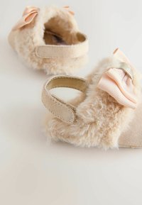 Chaussures pour tout-petits beiges avec texture en fausse fourrure, nœud décoratif rose et bande Velcro. Matériau doux et design arrondi pour le confort.