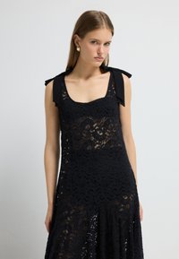 Sort blonde kjole med firkantet udskæring, skulderbuer og en udskåret kant. Det detaljerede blonde mønster tilføjer tekstur og detaljer.