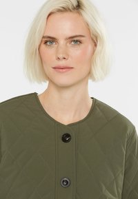 Blonde Frau mit kurzem Haar, die eine gesteppte olivgrüne Jacke mit großen schwarzen Knöpfen trägt, vor einem weißen Hintergrund.