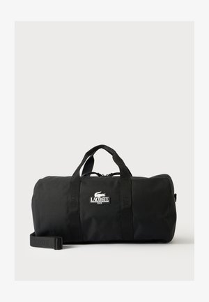 Borsa duffle nera Lacoste con doppio manico, tracolla e toppa con logo bianco sul davanti, su sfondo chiaro e semplice.