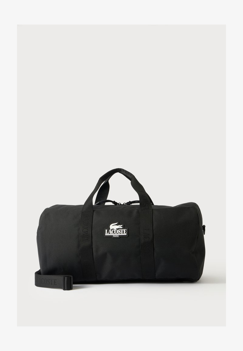Μαύρη τσάντα Lacoste τύπου duffle με διπλές λαβές, ιμάντα ώμου και λευκό σημάδι λογότυπου στο μπροστινό μέρος, τοποθετημένη σε απλό ανοιχτό φόντο.