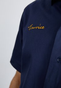 Navyblauwe korte mouw shirt van katoen met een knoopsluiting bij de kraag. "Service" geborduurd met gouden draad op de linkerkant van de borst.