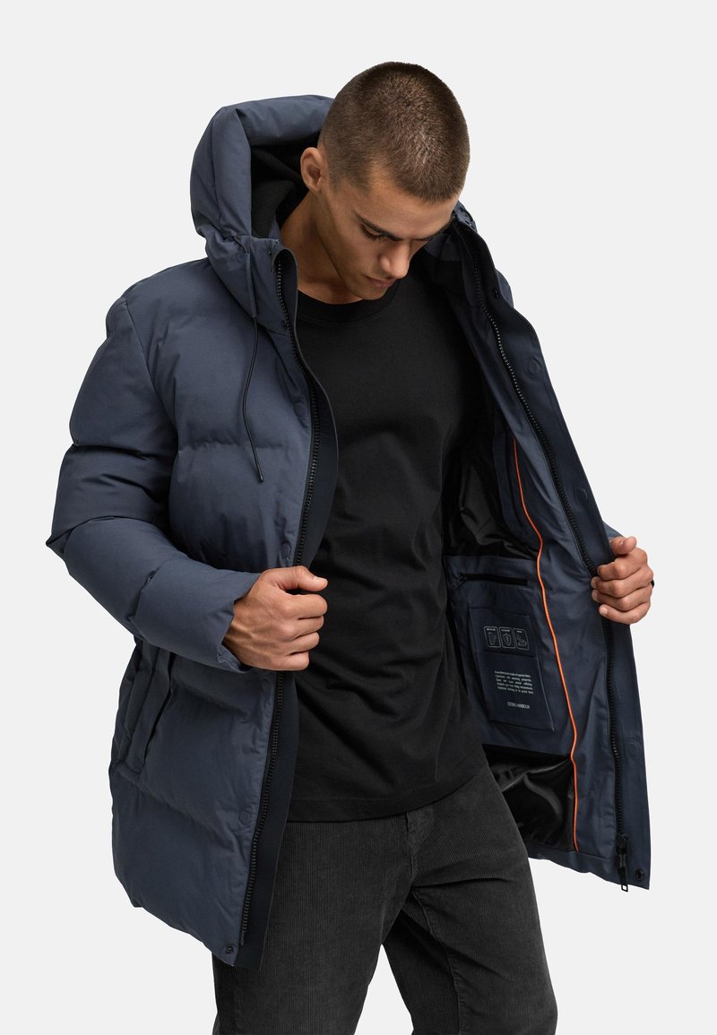 Temno modra pufasta jakna s kapuco, z zadrgo spredaj in elastičnimi manšetami. Ima notranji žep z oznakami in kontrastno oranžno podlogo.