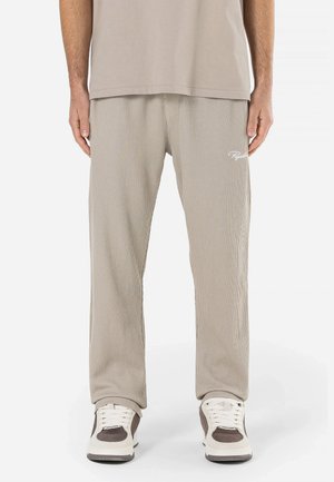 Beige ribbede sweatpants med en afslappet pasform, med et broderet logo på venstre lår og sammen med hvide og brune sneakers.