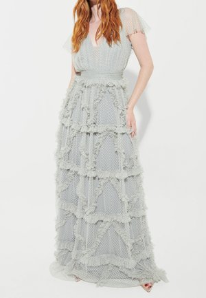 Woman with long red hair wearing floor-length pale blue dress with textured ruffles and sheer short sleeves, standing against white background.