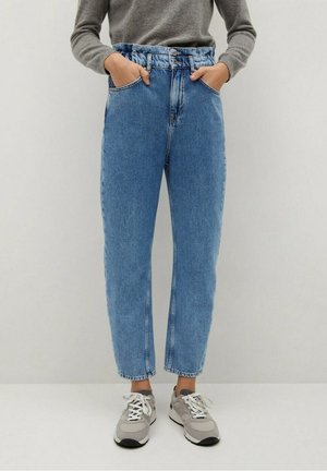 Personne portant un jean bleu taille haute avec jambes effilées, un pull gris et des baskets gris clair, les mains dans les poches avant.