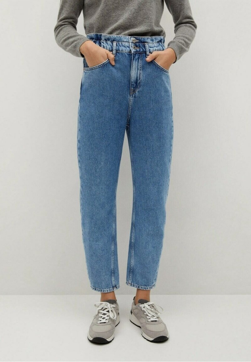 Personne portant un jean bleu taille haute avec jambes effilées, un pull gris et des baskets gris clair, les mains dans les poches avant.