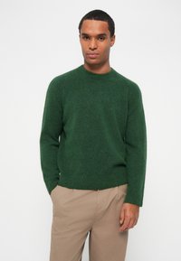Pulôver verde tricotado com decote redondo, mangas longas e barra canelada. O material parece suave e texturizado, usado com calças bege.