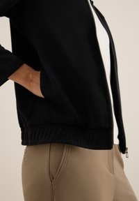 Veste zippée noire avec taille élastique, portée sur un haut blanc. Pantalon beige avec poches latérales et détails de couture visibles.