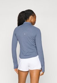 Langermet, lyseblå sportstrøye med høy hals, glatt tekstur og subtile logodetaljer på ryggen, kombinert med hvite shorts.