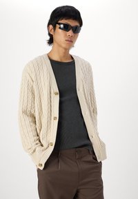 Kremfarget kabelstrikket kardigan med V-hals og fem knapper, iført over en mørkegrå teksturert skjorte, kombinert med brune bukser og svarte solbriller.