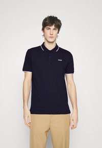 Marinblå piké med vit krage och ärmdekorer, med tre knappar och en liten vit logotyp på bröstet. Enfärgad texturerad tyg.