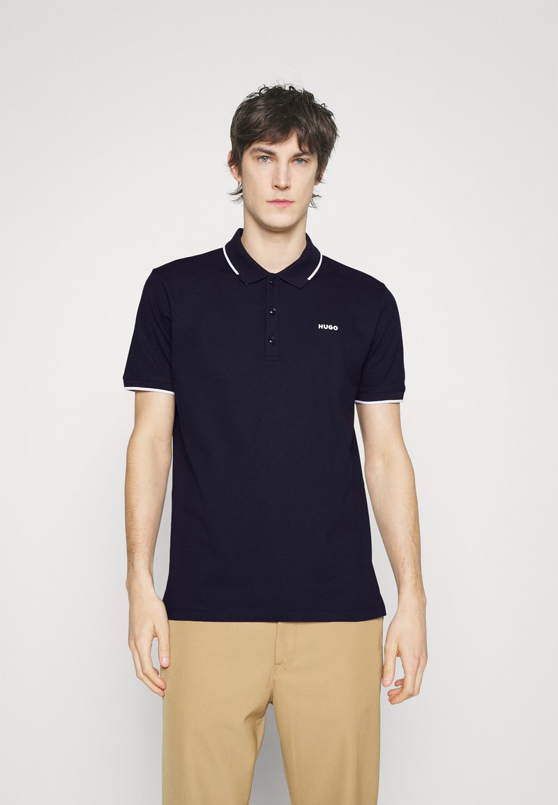 Marinblå piké med vit krage och ärmdekorer, med tre knappar och en liten vit logotyp på bröstet. Enfärgad texturerad tyg.