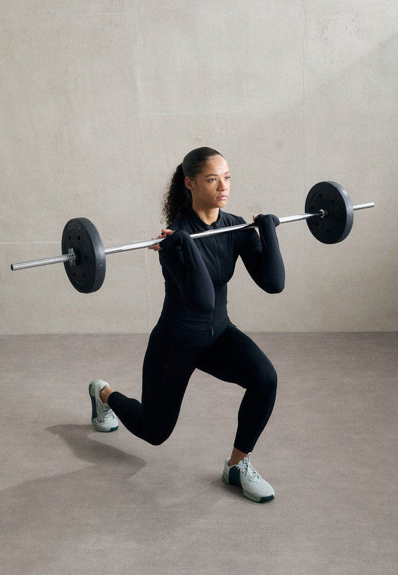 Barra de levantamiento de pesas con discos negros sostenida por una persona en una camiseta de manga larga negra ajustada y leggings negros, que lleva unos zapatos verde claro.