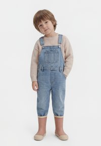 Copil tânăr cu păr scurt castaniu deschis, purtând un pulover bej, salopetă de denim cu mânecile suflecate și cizme de ploaie bej, stând cu o mână în buzunar.