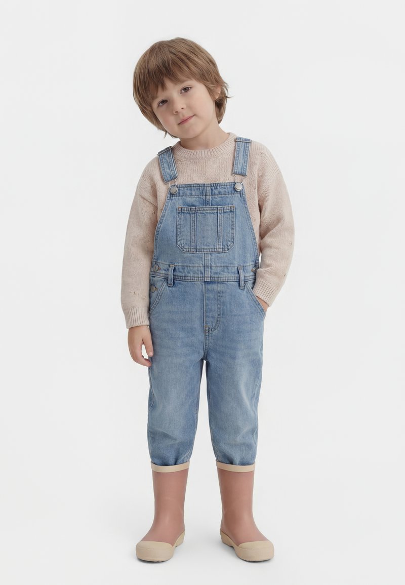 Copil tânăr cu păr scurt castaniu deschis, purtând un pulover bej, salopetă de denim cu mânecile suflecate și cizme de ploaie bej, stând cu o mână în buzunar.