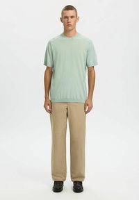 T-shirt en maille à manches courtes vert clair associé à un pantalon beige à jambes droites et des chaussures noires, debout sur un fond blanc uni.