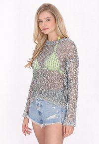 Sidabrinė permatoma megztinė su stačiakampiu raštu, ilgos rankovės ir apvalus iškirpimas, uždėta ant ryškiai žalio maudymosi kostiumėlio viršaus ir džinsinių šortų.