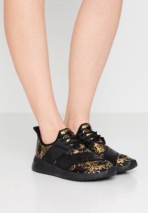 Jalat, joissa on mustakultakuviolliset tennarit mustilla nauhoilla ja joustavilla hihnoilla, vaaleanharmaalla lattialla.