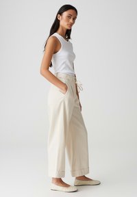 Crème-kleurige wijde broeken met een verstelbare tailleband, zijzakken en omslag aan de broekspijpen, gecombineerd met een strakke witte tanktop en platte schoenen.