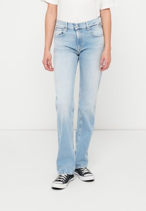 Világoskék farmernadrág, egyenes szárú fazonnal, öt zsebbel és gombos zipzárral. Fekete vászon sportcipővel viselve.