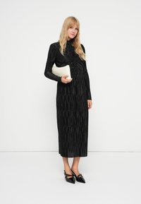 Fekete, hosszú ujjú ruha texturált redőkkel, testhez simuló felsőrész és egyenes maxi szoknya. A modell fehér clutch-t tart és fekete hegyes orrú cipőt visel.