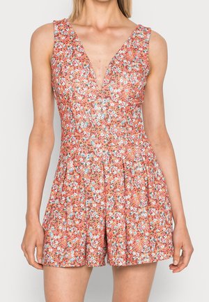 Mouwloze koraalkleurige romper met kleine witte, blauwe en gele bloemenprint en een diepe V-hals, gedragen door een staande vrouw.