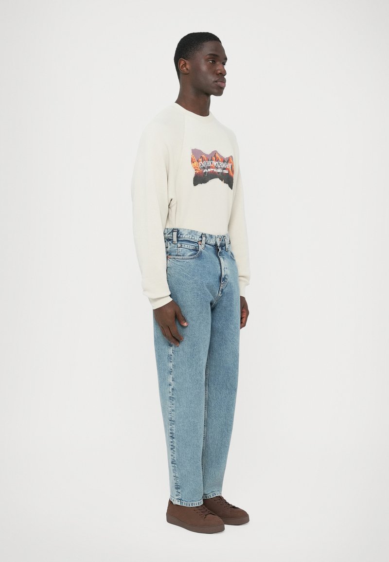 Light beige sweatshirt with a mountain graphic, paired with light blue denim jeans. Brown shoes complete the outfit.