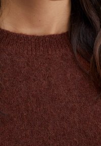 Pull en tricot marron avec un col côtelé. Texture douce avec des fibres visibles. Vue en gros plan se concentre sur la zone du col.