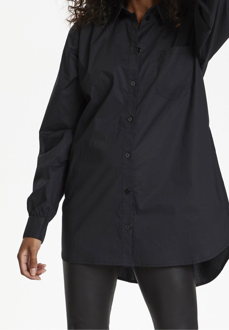 Femme portant une chemise noire à manches longues boutonnée avec un pantalon en cuir, montrée du cou jusqu'à mi-cuisse sur fond blanc.