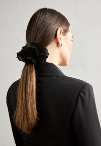 Juodas nėrinių scrunchie su sluoksniuotais volanais, dėvimas ilgose rudose plaukų sruogose, surištuose atgal, kontrastuojantis su juodu švarku.