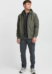 Maslinasto zelena bomber jakna s kapuljačom, uskim manžetama i prednjim patentnim zatvaračem. Uparena s tamnosivim cargo hlačama i svijetlosivim tenisicama.