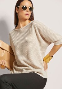 Beige gebreide trui met een textuurpatroon, brede halslijn en verlaagde schouders, gecombineerd met zwarte broek en een beige handtas. Accenten zijn onder andere een paar zonnebrillen en een gouden armband.