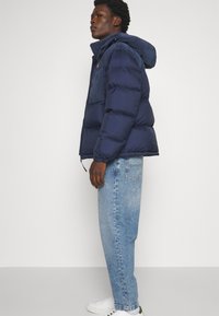 Giacca puffer blu navy con cappuccio, caratterizzata da un pannello in velluto a coste, zip laterali e una patch con logo. Indossata con jeans azzurri e sneakers bianche.