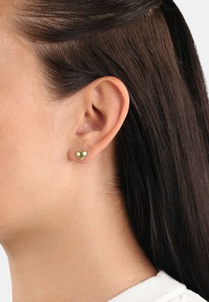 Pendiente de stud en forma de corazón de oro con superficie pulida, colocado en una oreja perforada. El pelo está peinado de forma suave, pegado al rostro, mostrando el pendiente.