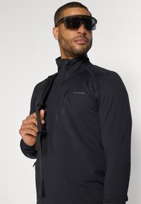 Zwarte lange mouwen pullover met een halve ritssluiting kraag, gestructureerde stof en een klein ritszakje op de borst. Inclusief verstelbare strap.
