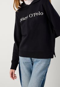 Czarna bluza z kapturem, wykonana z bawełny, z białym logo "Marc O'Polo" z przodu, kangurzą kieszenią i ściągaczami na rękawach.