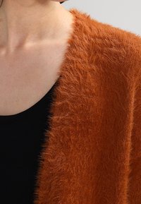 Rostfärgad, fluffig kofta med en mjuk textur, bärs över en svart topp. Designen har en öppen framsida och en avslappnad passform.