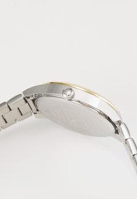 Reloj de pulsera con tonos plateado y dorado, con una caja redonda, banda de metal texturizada y una corona lateral para ajuste. Presenta un acabado suave.