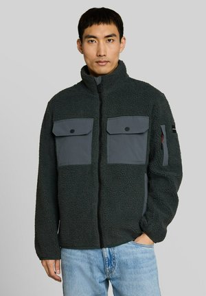 Uomo che indossa una giacca in pile verde scuro con due tasche grigie sul petto e jeans blu, in piedi davanti a uno sfondo chiaro e uniforme.