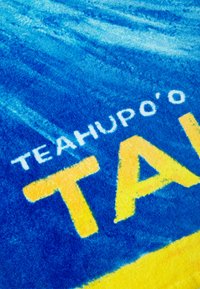 Sininen ja keltainen pyyhe, jossa on valkoisella teksti "TEAHUPO'O" ja keltaisella "TAL", pehmeä ja teksturoitu kangas.